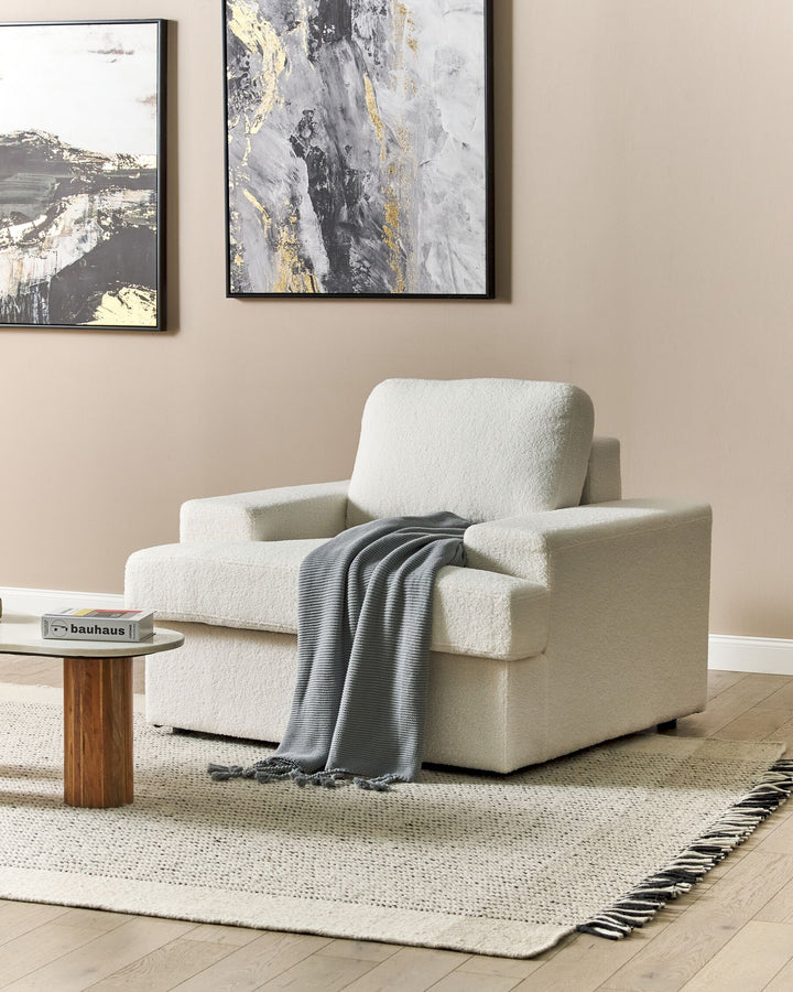 An off-white upholstered armchair with a wooden stable frame, decorated with a grey blanket, placed in a cozy living room setting with a modern abstract painting on the wall.