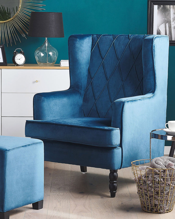 A dark blue velvet wingback armchair with a square footstool, situated in a room with a teal wall and a variety of decor.