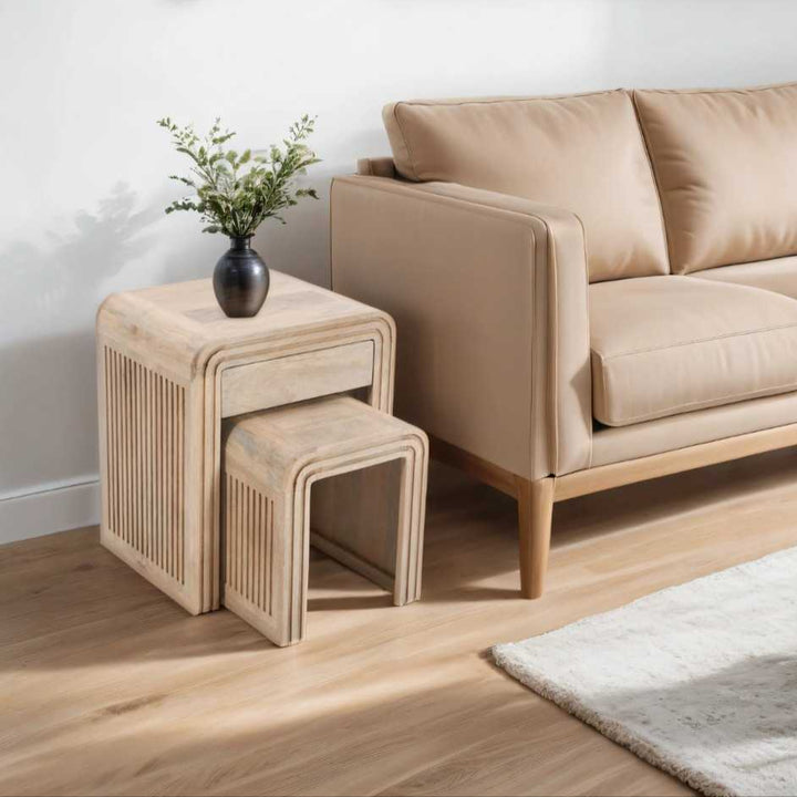 Nest of two carved mango wood tables with a drawer, placed beside a sofa on a wooden floor, with a potted plant on top of one of the tables.
