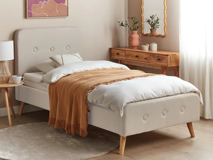 Beige upholstered bed with white bedding and a brown throw blanket in a bedroom setting.