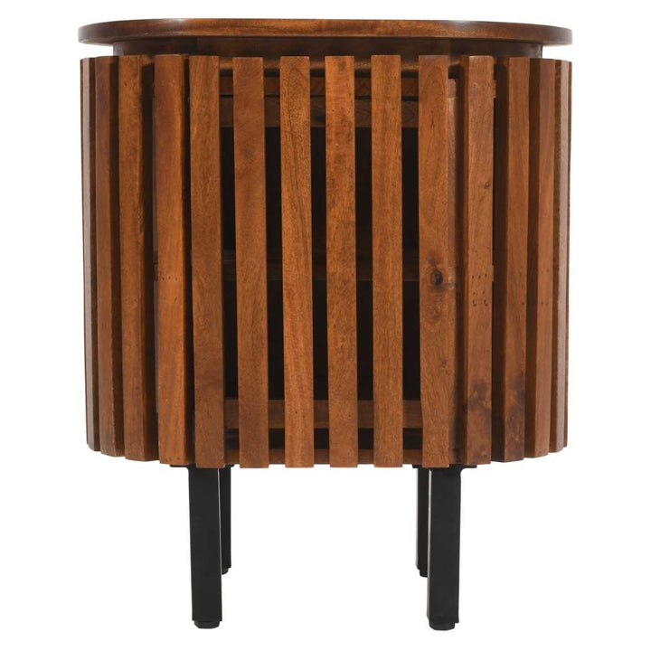 A round, dark mango wood bedside table with slatted side panels and a black front leg, with a door on the front for storage.
