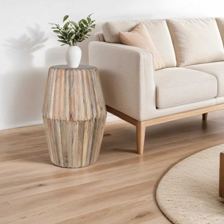 A carved solid mango wood round side table with a natural lacquer finish, displayed in a living room setting with a plant on top.