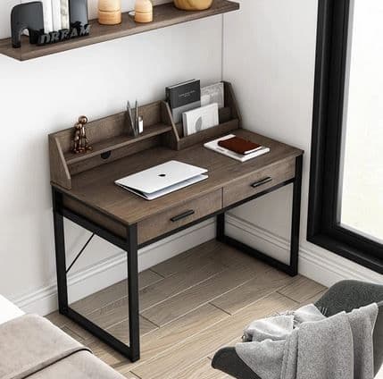 A corner desk with a brown wood-grain top and backrest, black metal legs, and two front drawers, shown in a home office setting with a laptop, books, and a shelf above.
