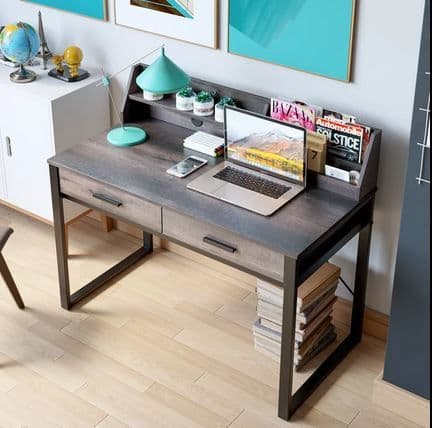A modern gray wood-grain writing desk with dark metal legs, styled with a laptop, lamp, and books in a home office setting.