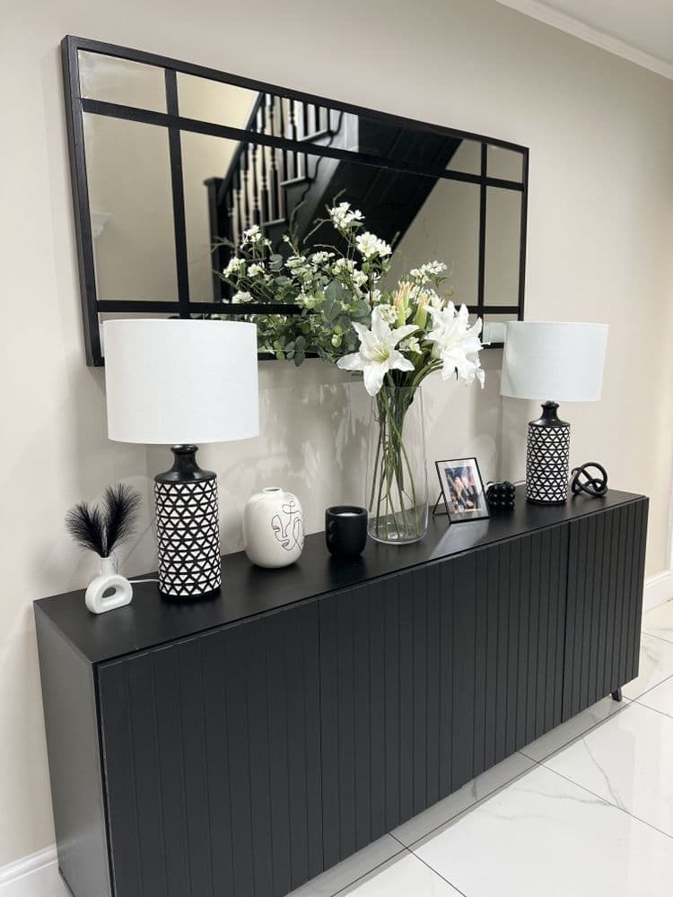 A long black sideboard or credenza in a home entryway, styled with two matching black-and-white patterned table lamps, white flowers in a glass vase, small decorative objects, and a large black-framed mirror on the wall above.