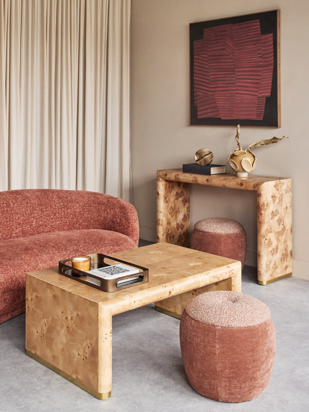 A styled interior scene showing a light natural burl wood coffee table in the foreground, a matching burl wood console table against the wall, two dusty rose velvet poufs, and a curved dusty rose velvet sofa on the left, with a framed abstract artwork on the beige wall.