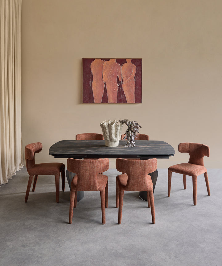 A dining set consisting of a dark rectangular table and six upholstered chairs in a warm rust-brown fabric, arranged in a minimalist dining room with a beige wall, a painting, and a gray floor.