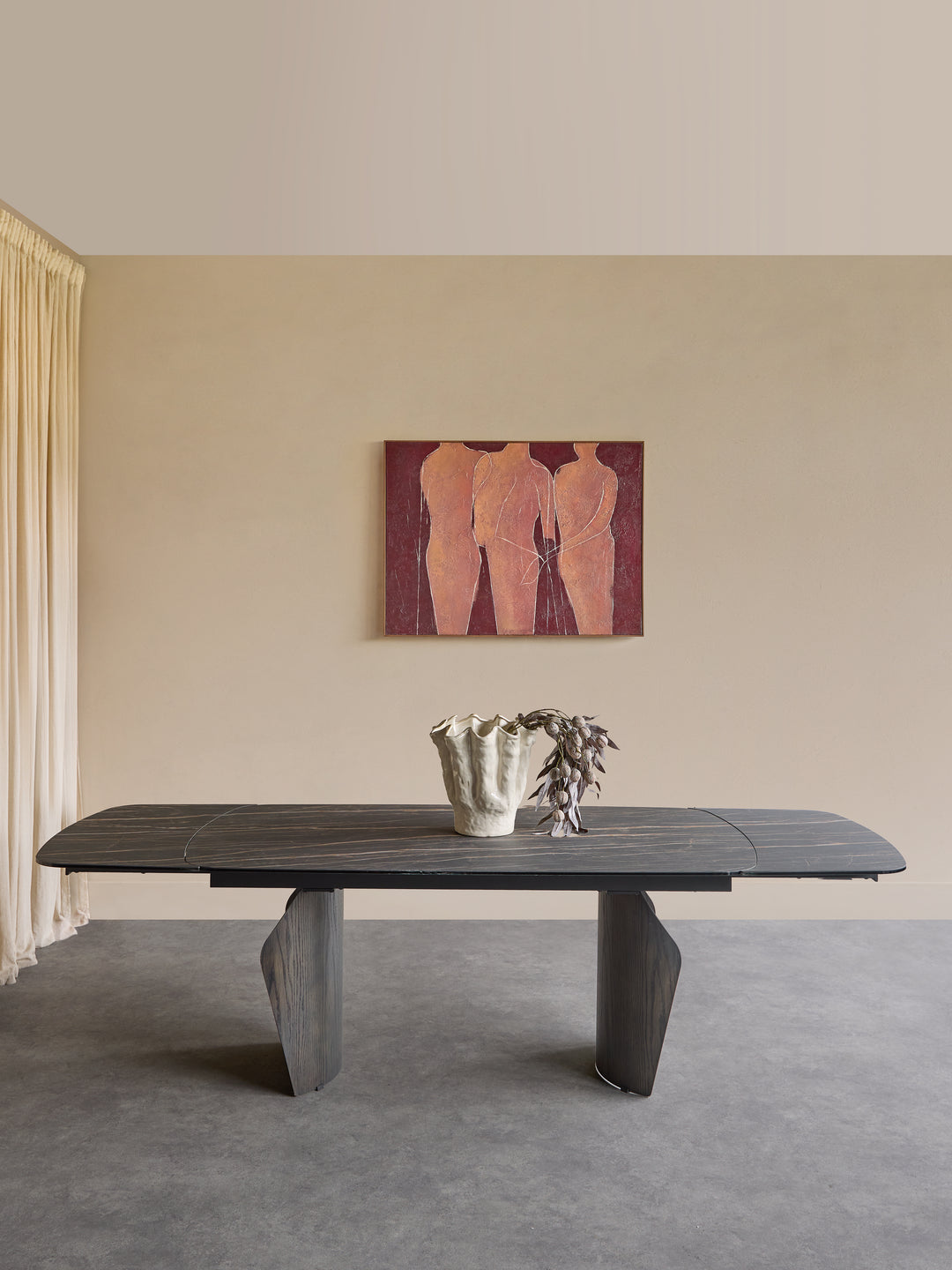 A dark wood dining table with sculptural gray legs is set in a minimalist room with a beige wall, a framed painting of three figures, and a small vase on top.