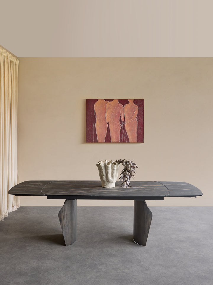 A dark wood dining table with sculptural gray legs is set in a minimalist room with a beige wall, a framed painting of three figures, and a small vase on top.