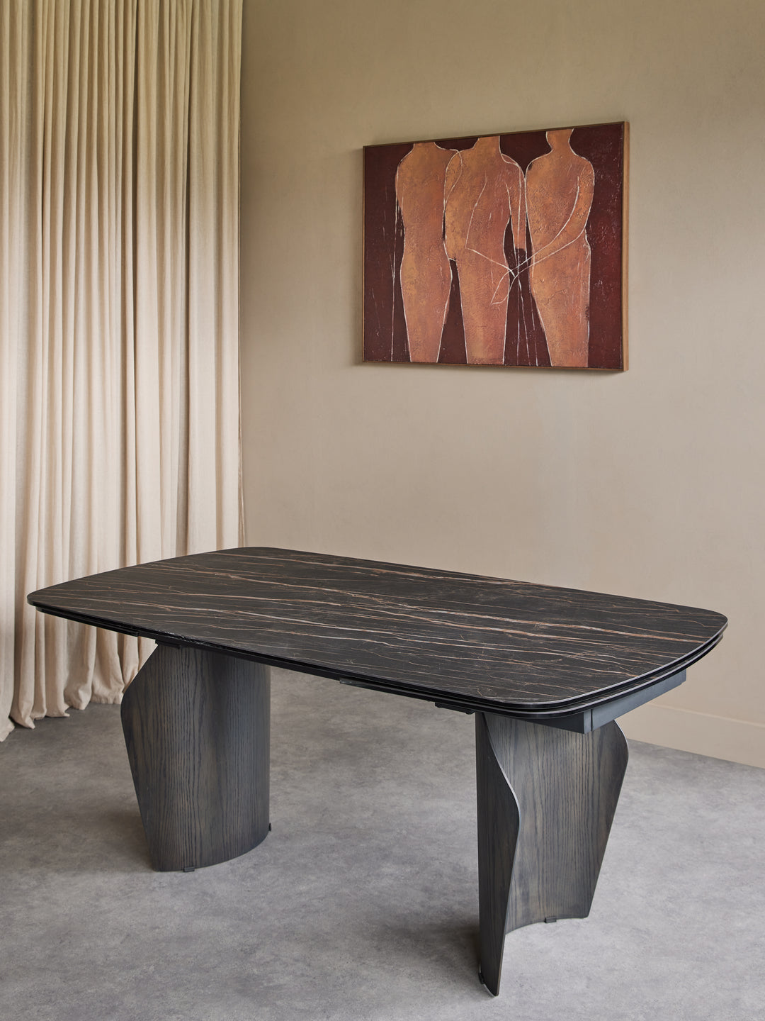 A dark wood dining table with sculptural, tapered legs and a rounded rectangular top, set in a modern room with beige walls, draped curtains, and a framed abstract painting.