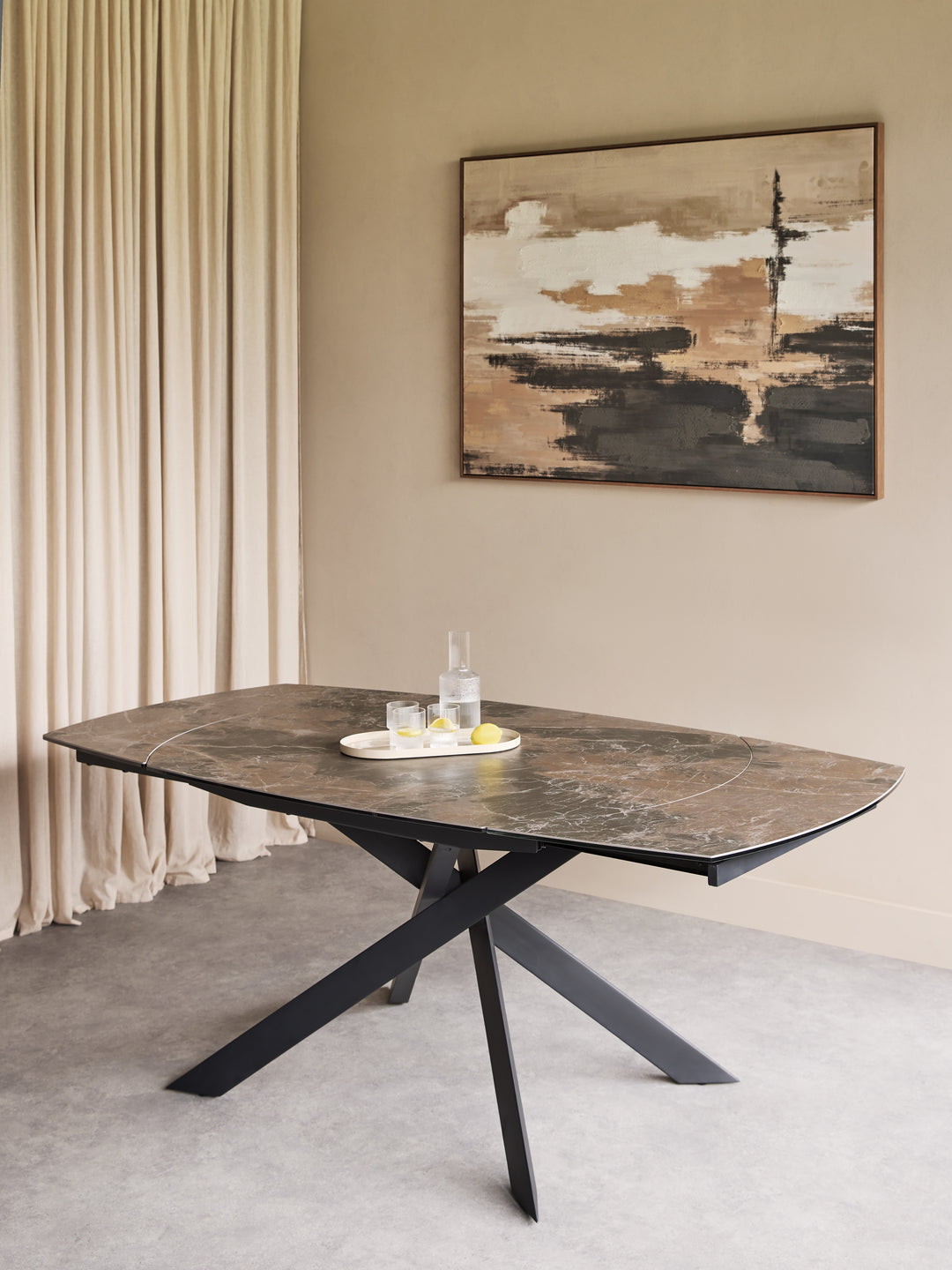 A modern dining table with a dark brown marble-look top and a black metal X-shaped base, styled in a minimalist room with beige walls, cream curtains, and a framed abstract painting.
