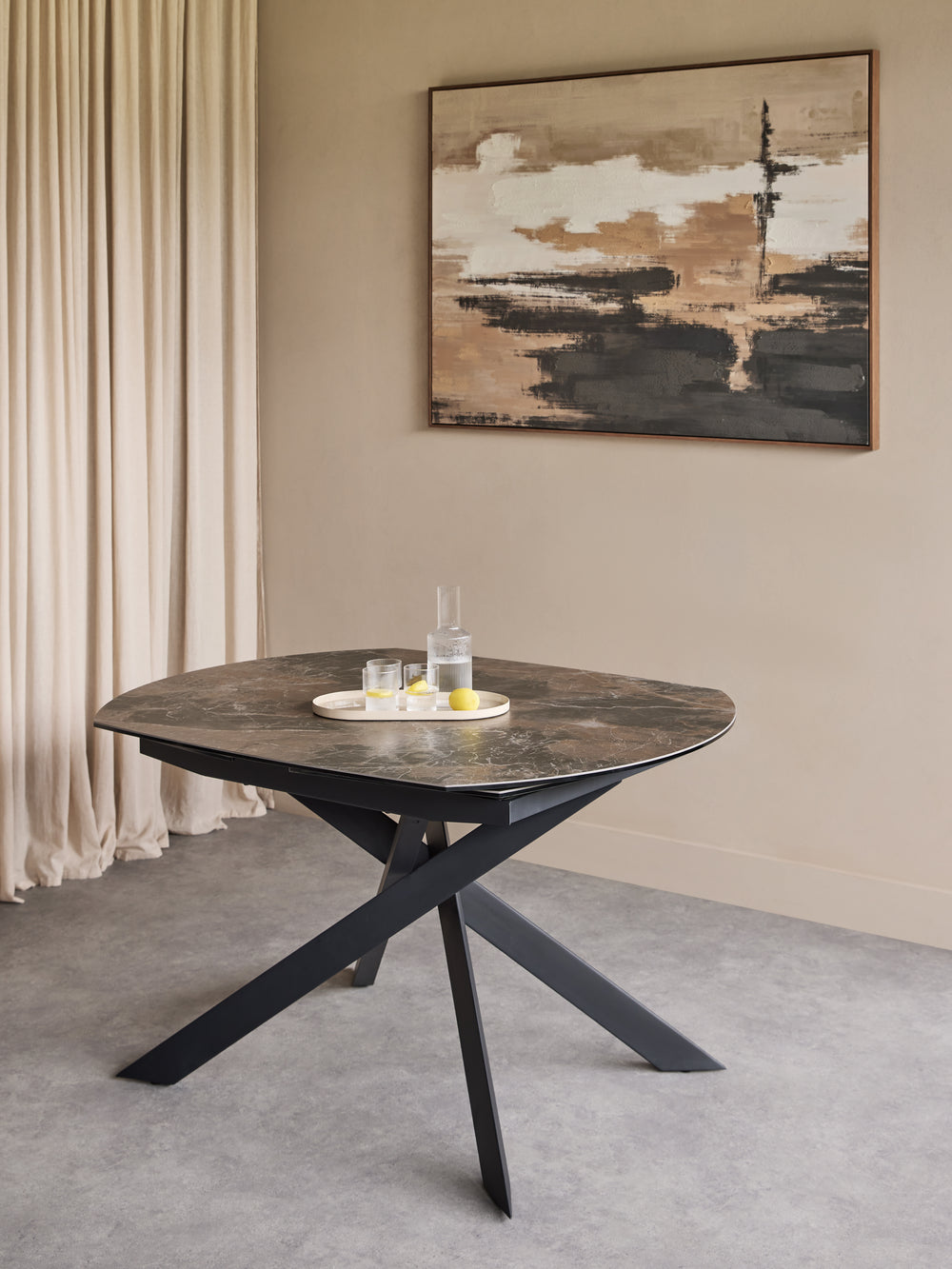 A modern round dining table with a dark stone-look top and black metal X-shaped legs, styled in a minimalist room with a beige wall, abstract painting, and cream curtains.