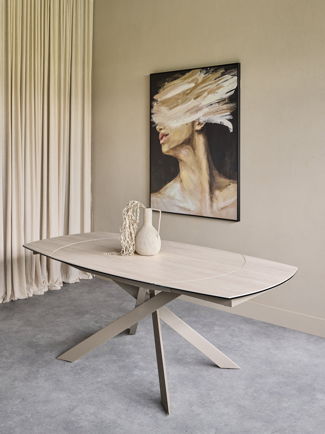 A modern oval dining table with a light wood top and angled metal legs, styled in a minimalist dining room setting with a framed painting on the wall and neutral decor.
