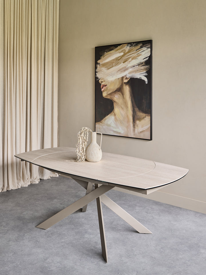 A modern oval dining table with a light wood top and angled metal legs, styled in a minimalist dining room setting with a framed painting on the wall and neutral decor.
