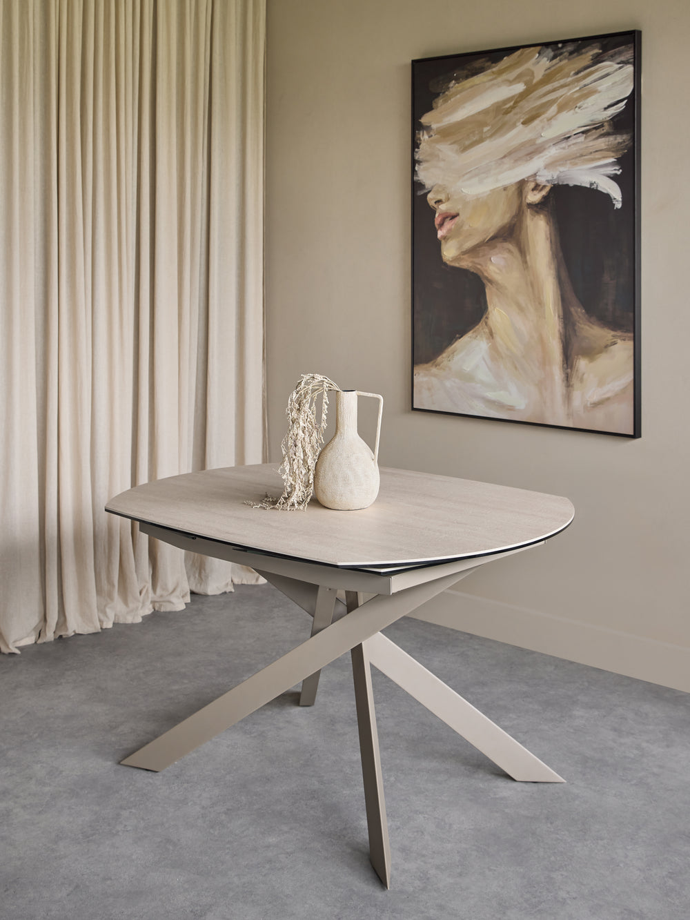 A light wood oval dining table with a sculptural cross base, styled in a minimalist room with beige curtains, a gray floor, and a framed abstract portrait on the wall.