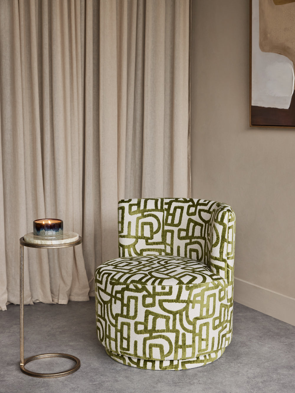 A modern barrel-style accent chair upholstered in an olive green and white geometric maze pattern, set in a styled room next to a small side table with a candle.