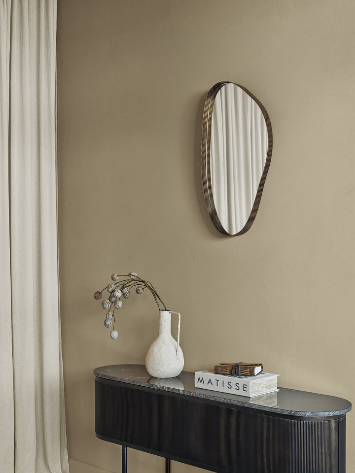 Round mirror with wooden frame on a beige wall above a dark console table with a vase and books.