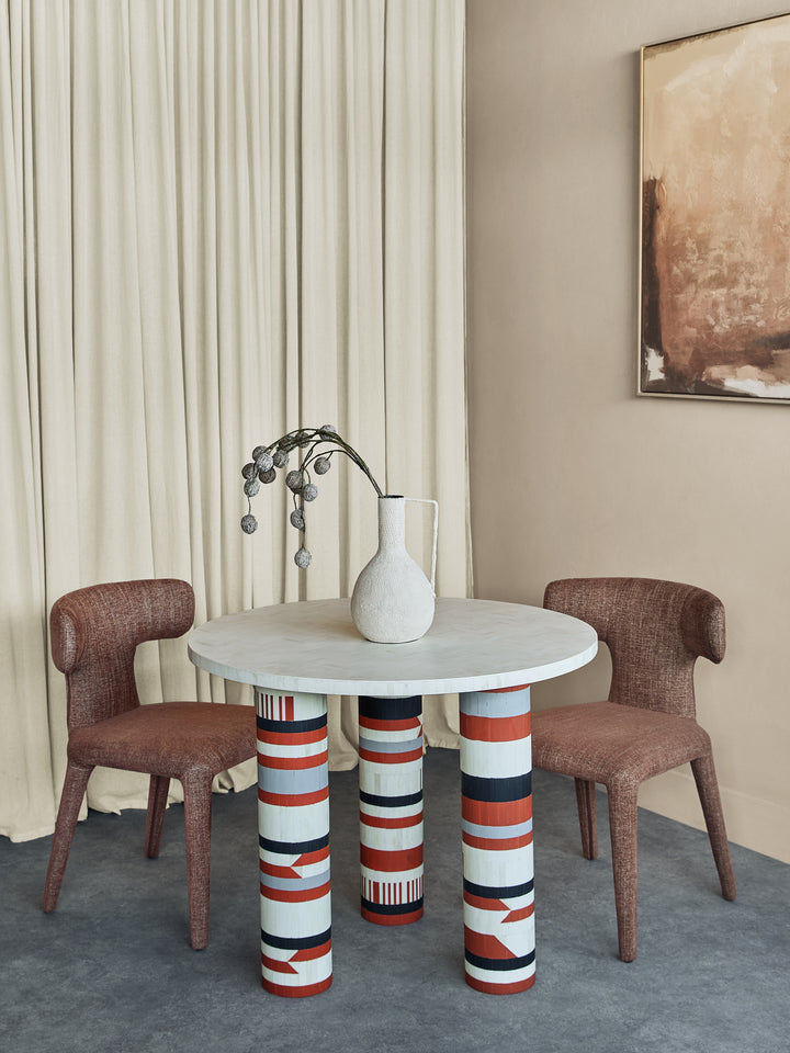 Round table with striped legs and two chairs in a room with curtains and a painting.