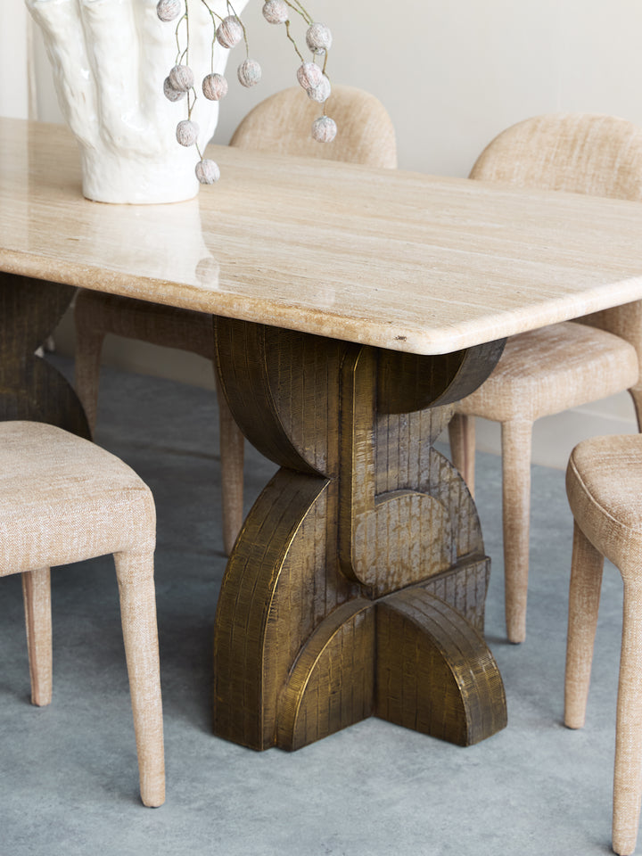 A dining table with a polished beige stone top and a sculptural dark wood pedestal base, surrounded by upholstered dining chairs in a neutral indoor setting.