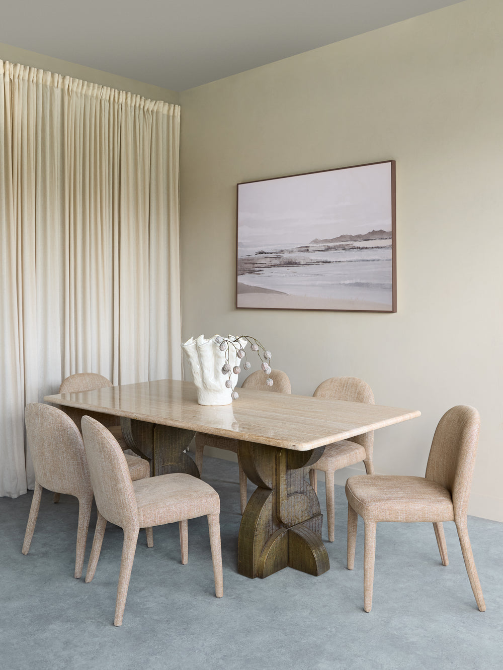 A modern dining room setup featuring a rectangular light wood table with a sculptural dark wood base, surrounded by six upholstered dining chairs in a neutral beige fabric. The room has beige walls, a blue-gray carpet, pleated cream curtains, and a framed landscape print on the wall.