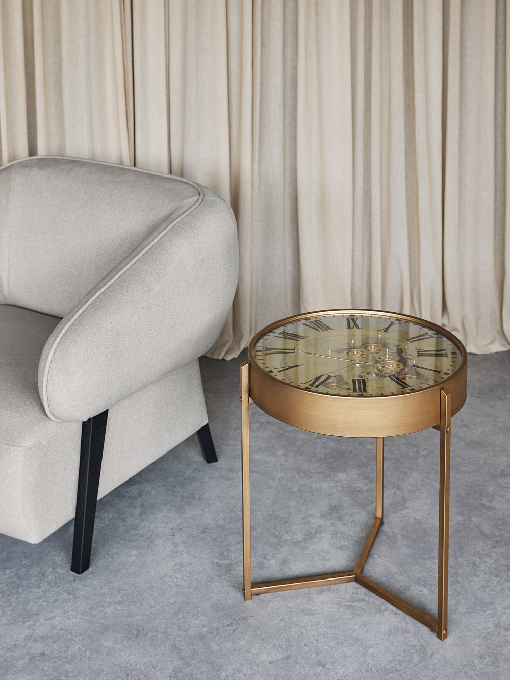 Gold side table with glass top next to a gray armchair in a room with beige curtains.