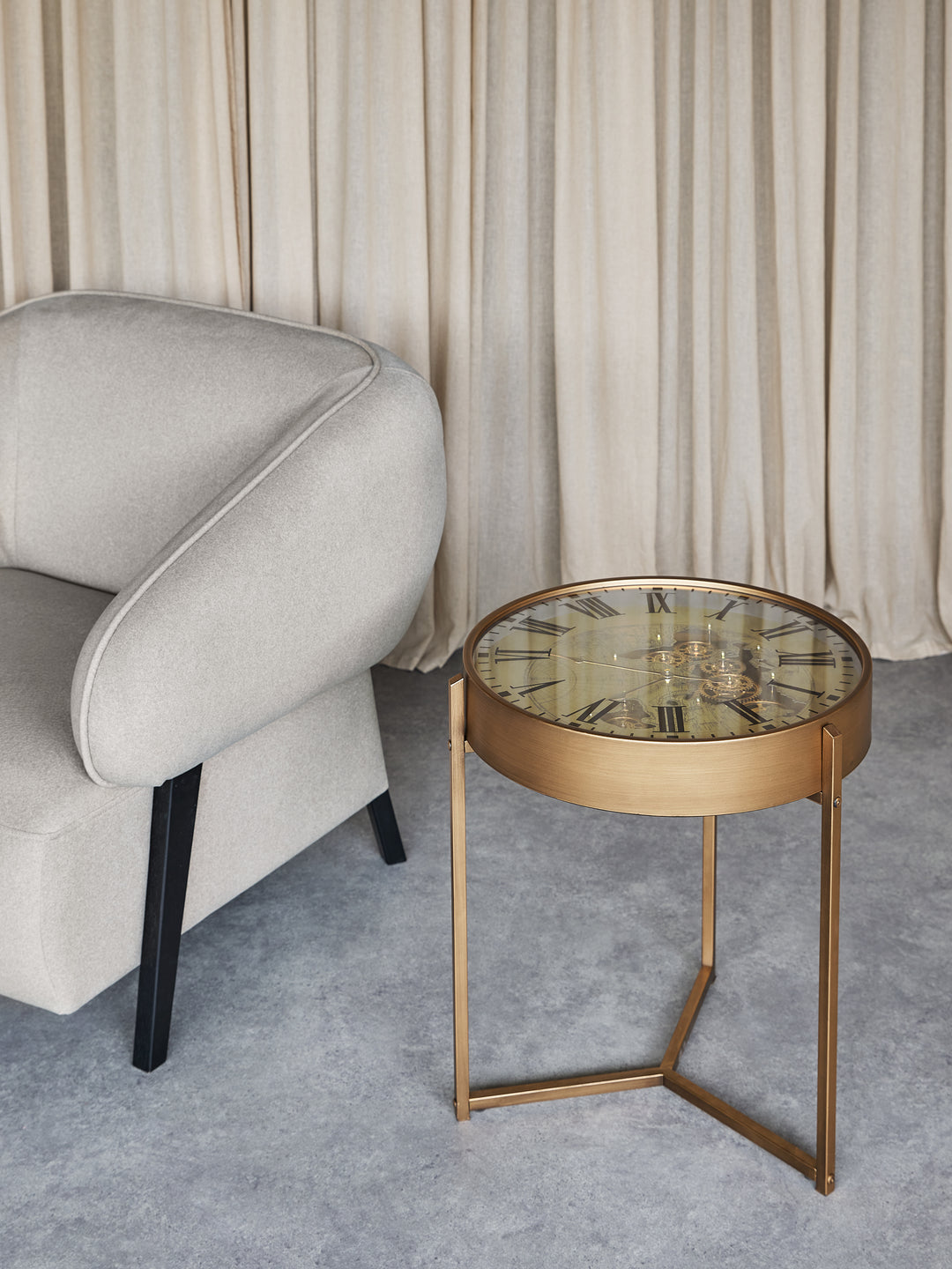 Gold side table with glass top next to a gray armchair in a room with beige curtains.