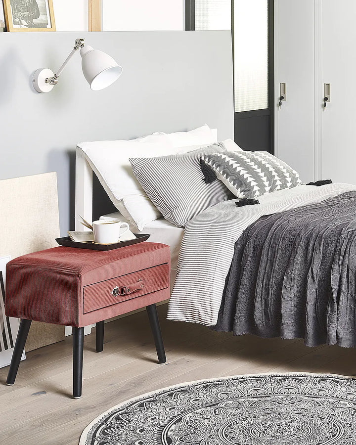 A styled bedroom scene featuring a bed with gray and white bedding, a dusty red upholstered nightstand with black legs, a white wall-mounted lamp, and a light wood floor.