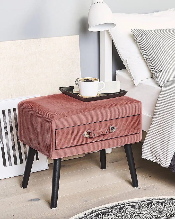 A dusty rose corduroy nightstand with black legs and a single front drawer, styled in a bedroom next to a bed with white linens and a lamp.
