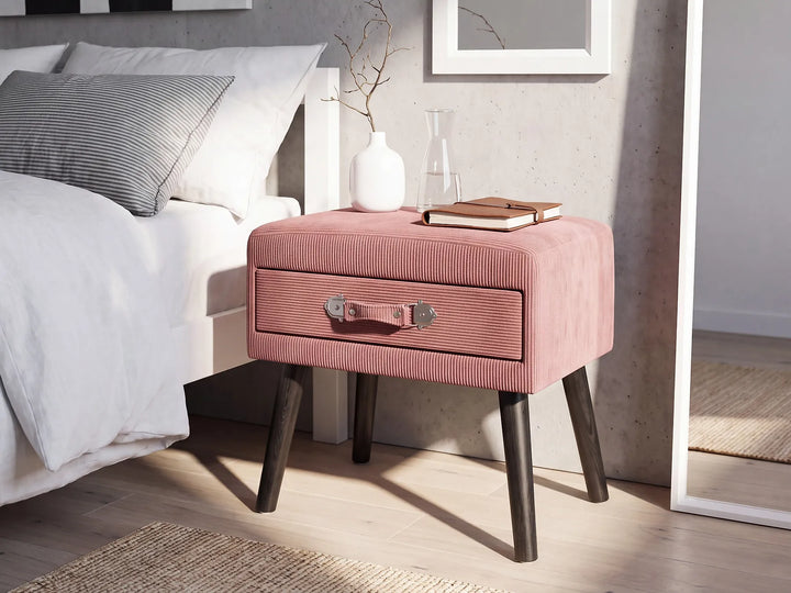 A pink corduroy nightstand with dark wood legs and a single front drawer, styled next to a bed in a modern bedroom setting.