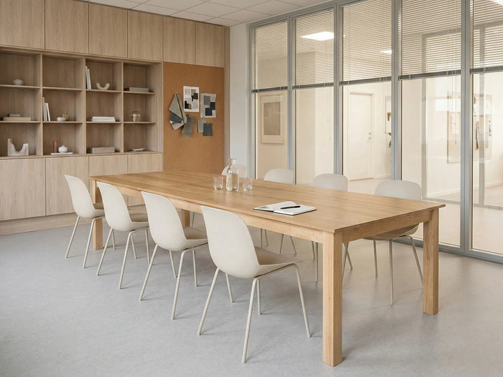 A modern meeting room featuring a light oak wood conference table surrounded by six white molded chairs with slim metal legs. The room has light wood cabinetry on the left, a cork board on the back wall, and large glass partition walls on the right.