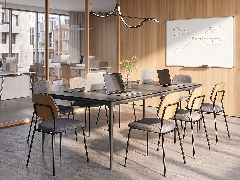 A modern conference room setup featuring a long rectangular black table surrounded by eight chairs with light wood and gray upholstery. The room has a wood-paneled wall with a whiteboard, and the table is styled with laptops and a small vase of greenery.