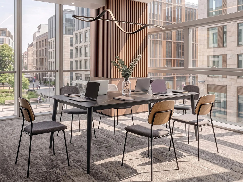 A modern meeting room setup featuring a dark rectangular conference table surrounded by six chairs with light wood backs and dark gray seats, set in a bright office with large windows overlooking city buildings.