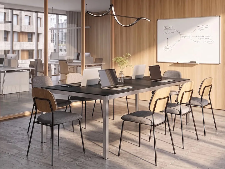A modern conference room setup featuring a long rectangular black table with a silver metal frame, surrounded by eight chairs with light wood backs and gray upholstered seats. Several laptops are open on the table, and a whiteboard is mounted on the wood-paneled wall in the background.
