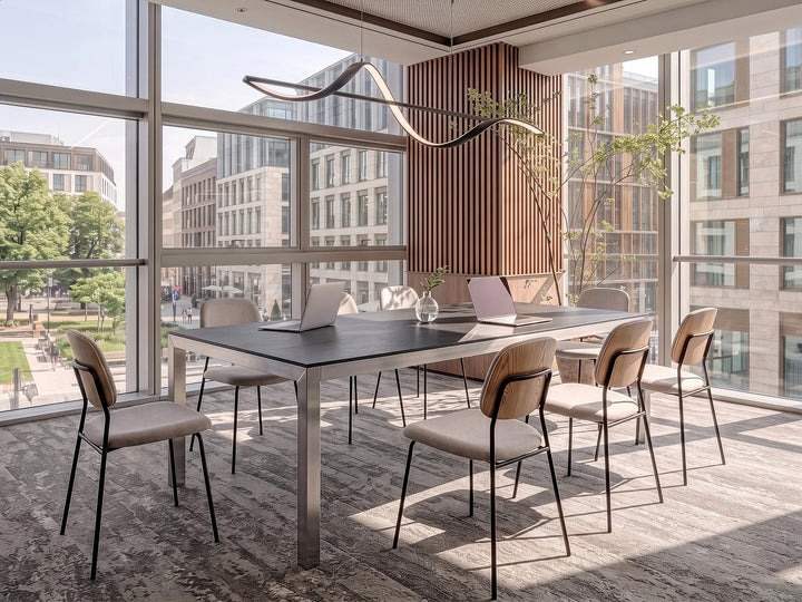 A modern meeting room setup featuring a rectangular dark gray conference table with a brushed metal base, surrounded by eight chairs with beige upholstered seats and light wood backs, set in a bright room with large windows overlooking a city park.