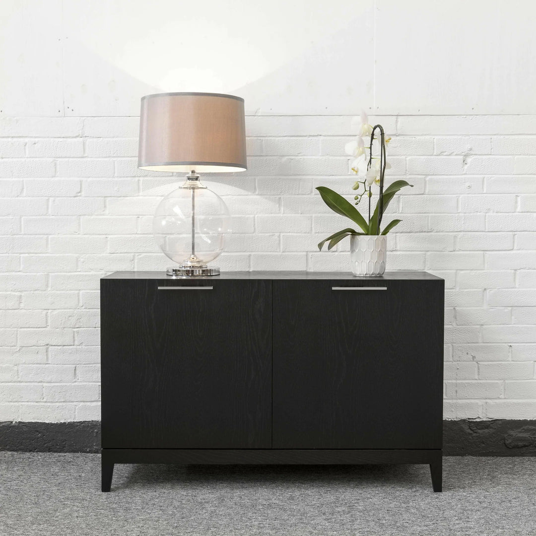 Black sideboard with a lamp and plant against a white brick wall.