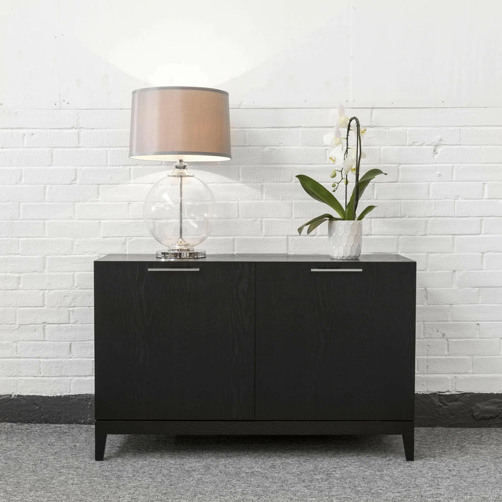 Black sideboard with a lamp and plant against a white brick wall.