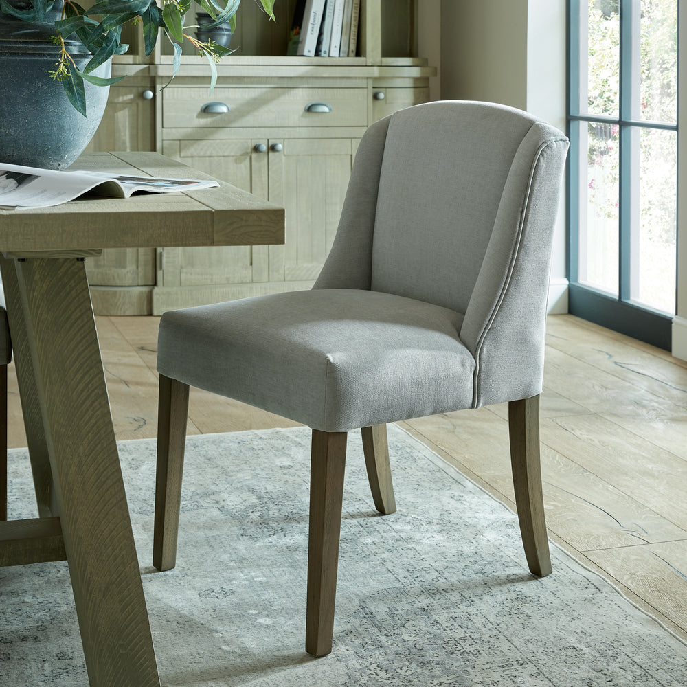 Gray upholstered chair next to a wooden table in a room with light wood flooring and a large window.