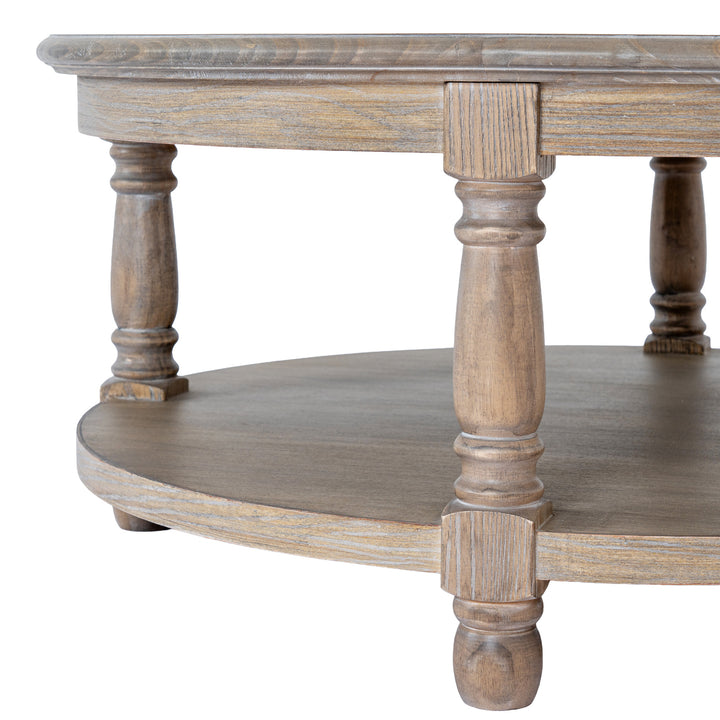 Close-up of a rustic round wooden coffee table with a distressed gray-brown finish, featuring a top surface, a lower shelf, and turned legs on a white background.