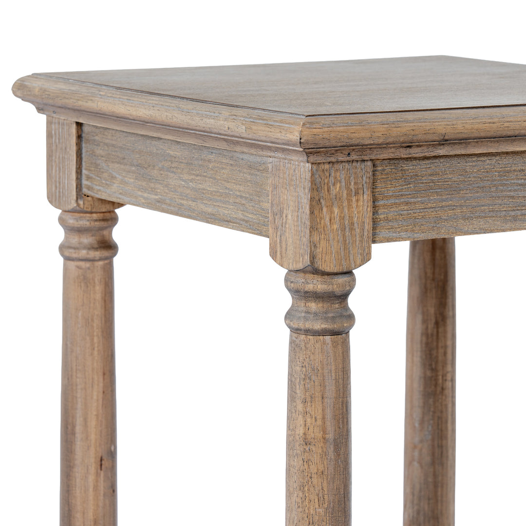 Close-up of a rustic wooden table corner showing the tabletop, apron, turned legs, and visible wood grain in a weathered brown finish against a white background.