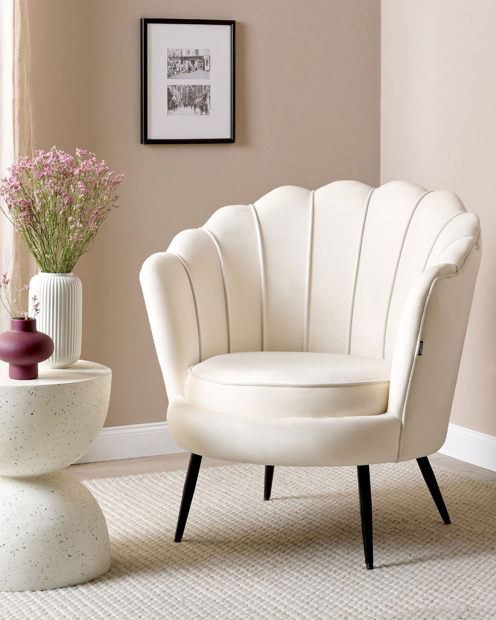 An off-white Lovikka armchair with metal legs and a plush velvet seat cushion, placed in a well-lit living room next to a round side table with a vase of pink tulips.