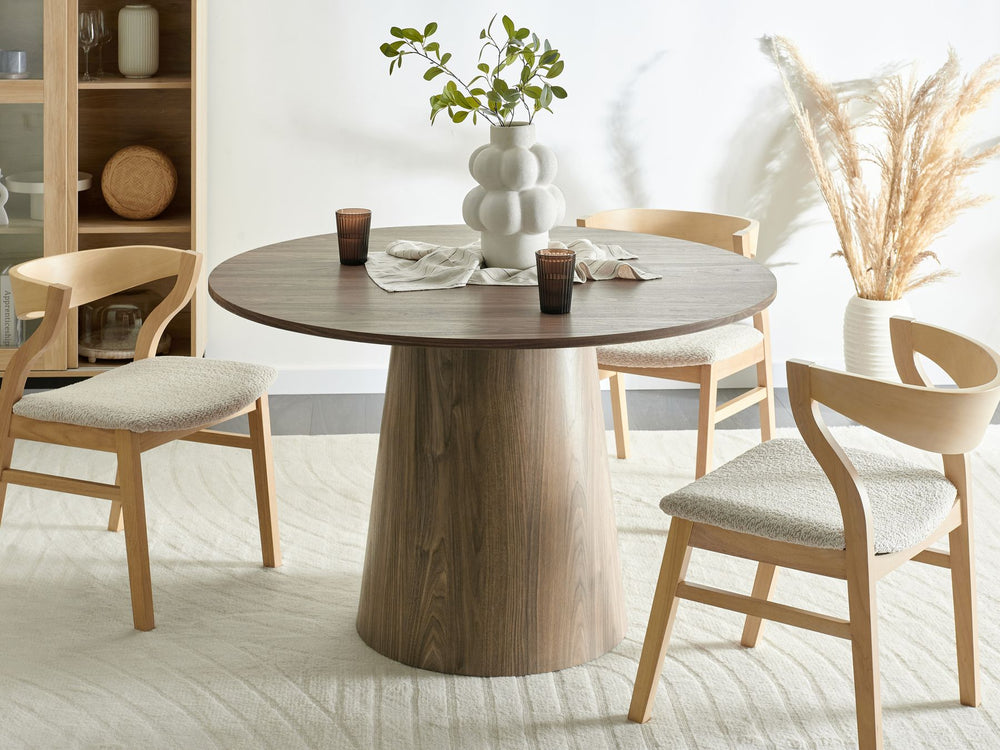 A round dining table in a room with light decor, featuring a dark wood finish and a pedestal base. The table is set with four chairs, and there is a vase with wheat in the background, indicating a cozy and inviting setting.