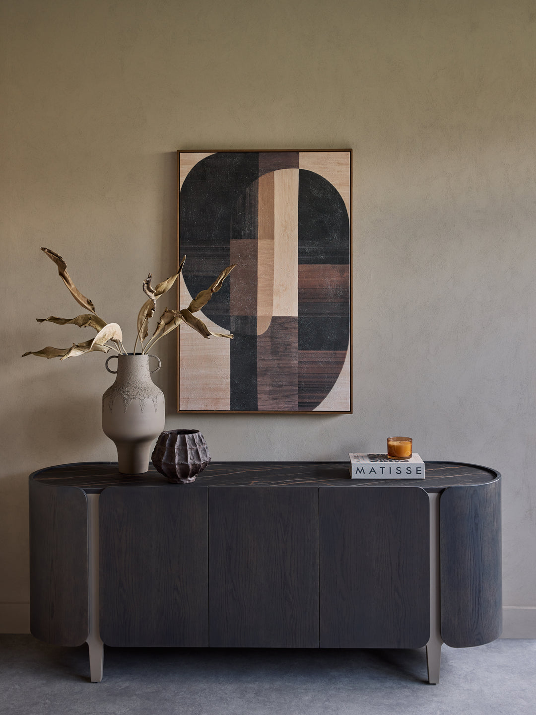 A dark wood console table in a minimalist room setting, styled with a ceramic vase containing dried branches, a small geometric sculpture, and a book. Above the table is a framed abstract geometric wall art piece.