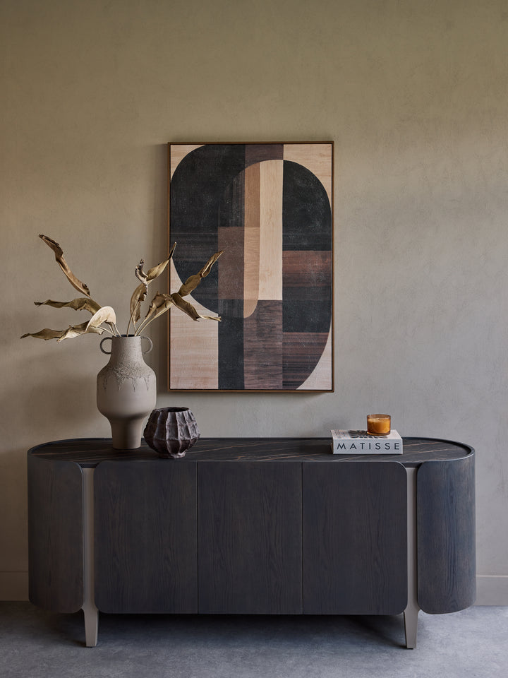 A dark wood console table in a minimalist room setting, styled with a ceramic vase containing dried branches, a small geometric sculpture, and a book. Above the table is a framed abstract geometric wall art piece.