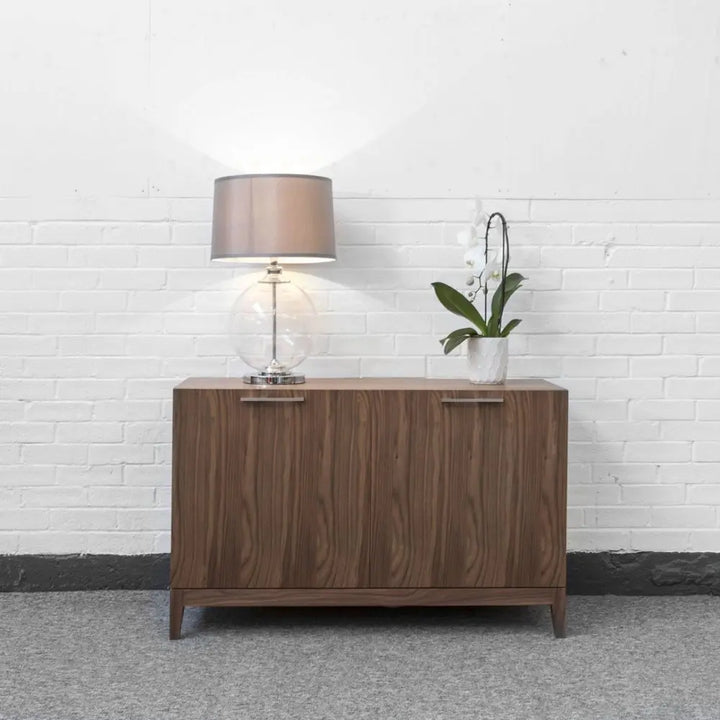 Wooden sideboard with a lamp and plant against a white brick wall.
