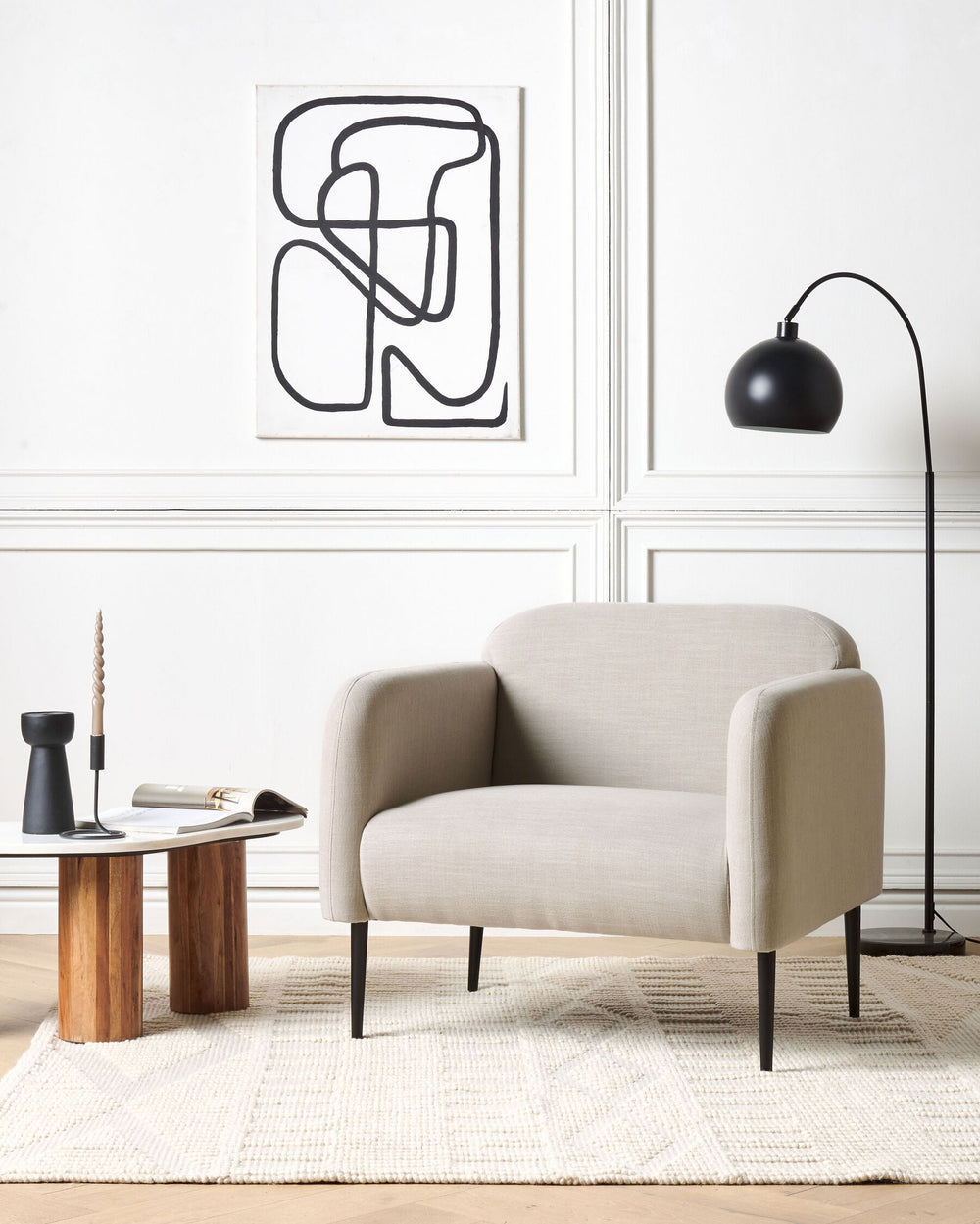 A taupe upholstered armchair with sleek metal legs, positioned in a well-lit living room next to a wooden side table with a lamp and a decorative item.