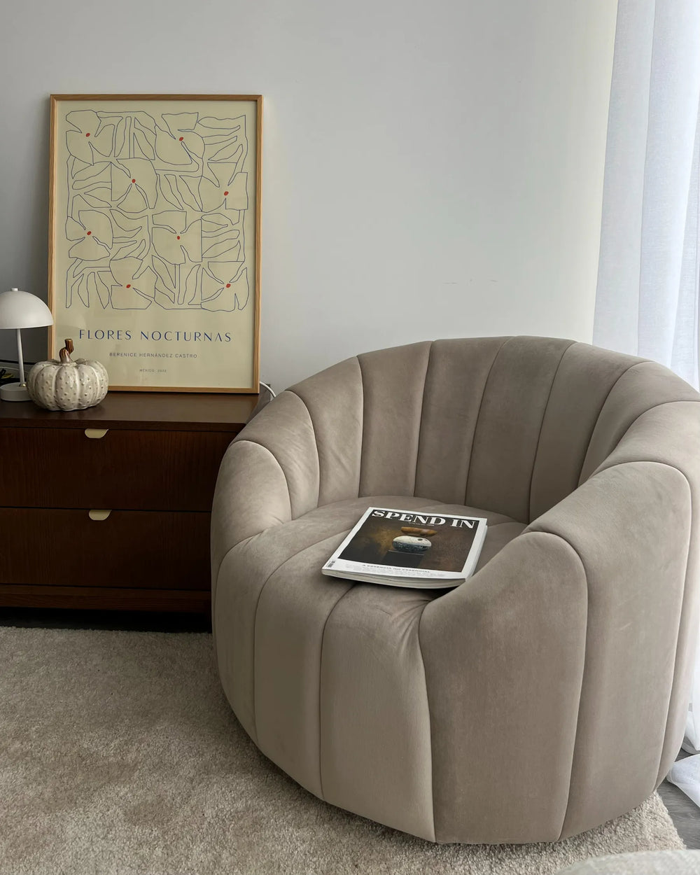 Beige armchair with a book on a carpeted floor, framed artwork on a wall.