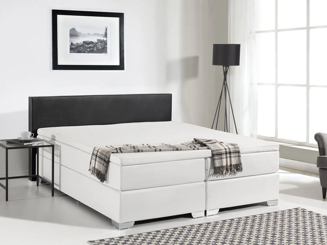 White bed with black headboard in a room with a lamp and picture on the wall.
