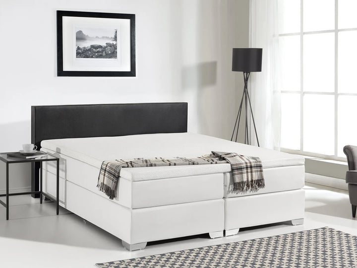 White bed with black headboard in a room with a lamp and picture on the wall.
