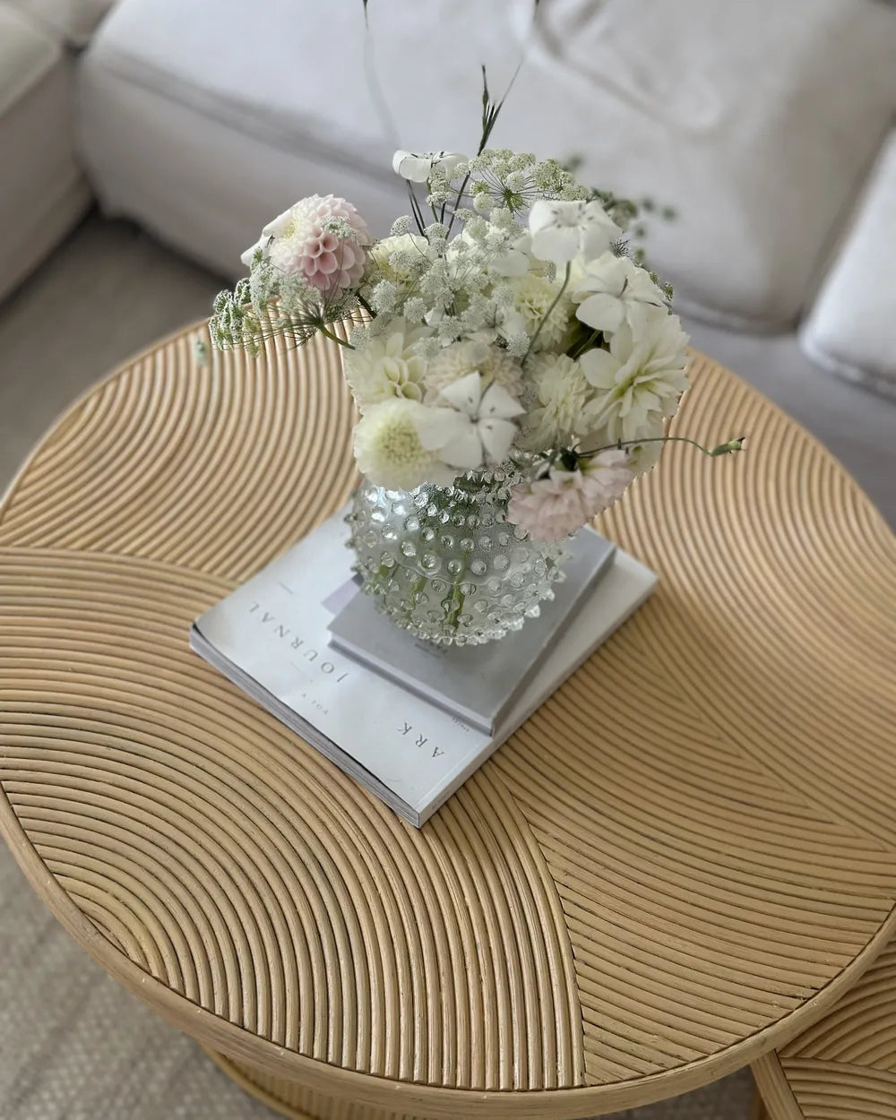 Round woven coffee table with a floral arrangement and books on top, in a living room setting.