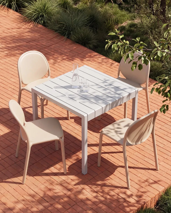 A square outdoor dining table with four matching chairs, set on a red brick patio with green plants in the background. The furniture has a light beige color with a white slatted tabletop.